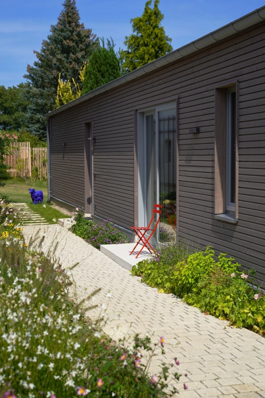 Ein modernes Gebäude mit einer Fassade aus dunkelbraunen Holzpaneelen. Ein roter Klappstuhl ziert das Podest der Eingangstür und ein naturbeige gepflasterter Weg im Reihenverband trennt das Gebäude von einem Garten im Vordergrund. Im Hintergrund sind einige Nadelbäume zu erkennen.