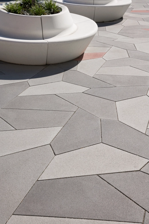 <span>fünfeckige Platten in verscheidenen Grautönen sind in in geometrischer Anordnung auf einem Platz verlegt. Darauf befinden sich Sitzbänke aus Sichtbeton, die im Kreis angeordnet und mittig bepflanzt sind.</span>