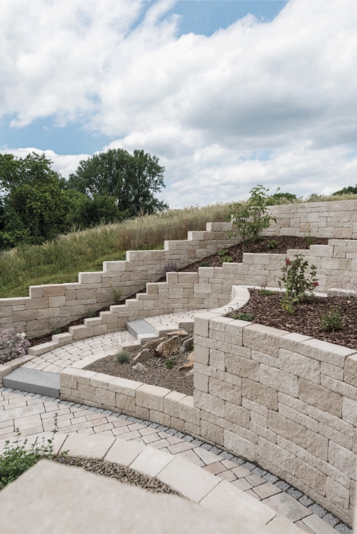 Mauersteine aus Beton in Kalksteinfarben und Naturstein-Optik 
terrassieren einen Hang in einem neu angelegten natürlichen Garten mit 
gemauerten PFlanzbeeten in unterscheidlcih abgestuften Höhen. ein 
geschwungener Weg mit sandgrauen Pflastersteinen verfläuft geschwungen 
zwischen abgetreppten Mauern.<br>