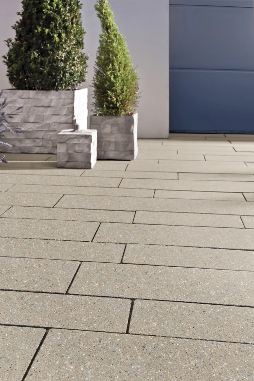 befahrbare Einfahrt mit beigen Terrazzo Pflastersteinen in 8 cm Dicke 
vor einem Garagentor in Anthrazit. Auf der Fläche stehen graue 
Pflanzkübel und ein Windlicht.