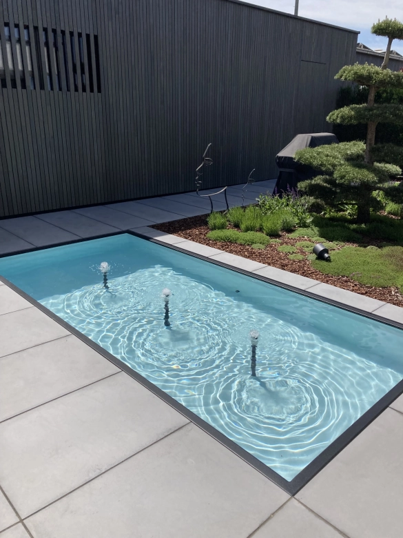Wasserbecken mit Fontäne vor einen mit Holz verkleidetes Gebäude und großen Terrassenplatten