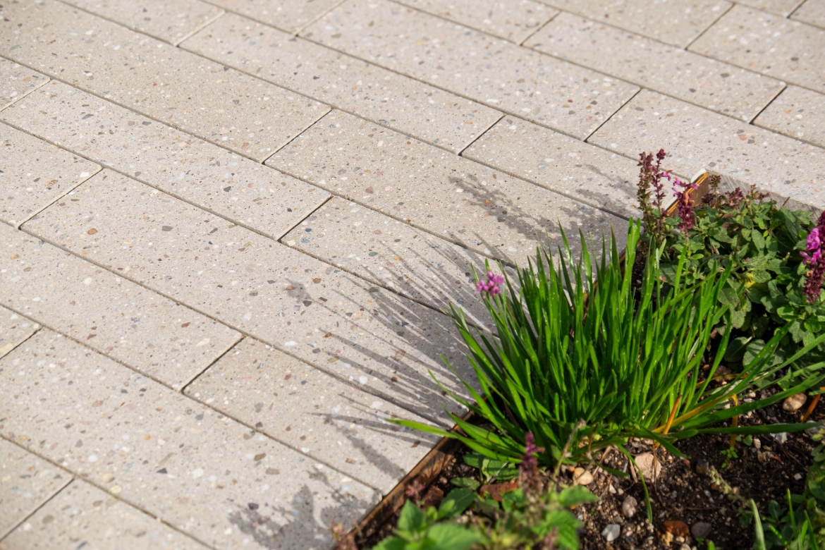 sandgraue Terrazzo Steine mit sichtbarer Splittkörnung in länglichem 
Format verlegt in versetztem Reihenverband. Eingefasst  mit einer 
Metallschiene aus Cortenstahl in Rostoptik.