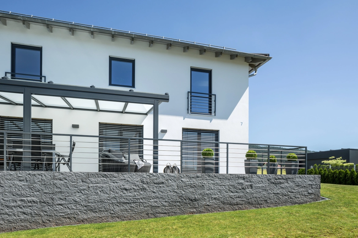 modernes weißes Haus mit erhöhter Terrasse und gemeuarter Stützwand in Anthrazit. Terrassenüberdachung aus Metall mit Glasdach in Anthrazit. Davor grüner, gepflegter Rasen .<br>