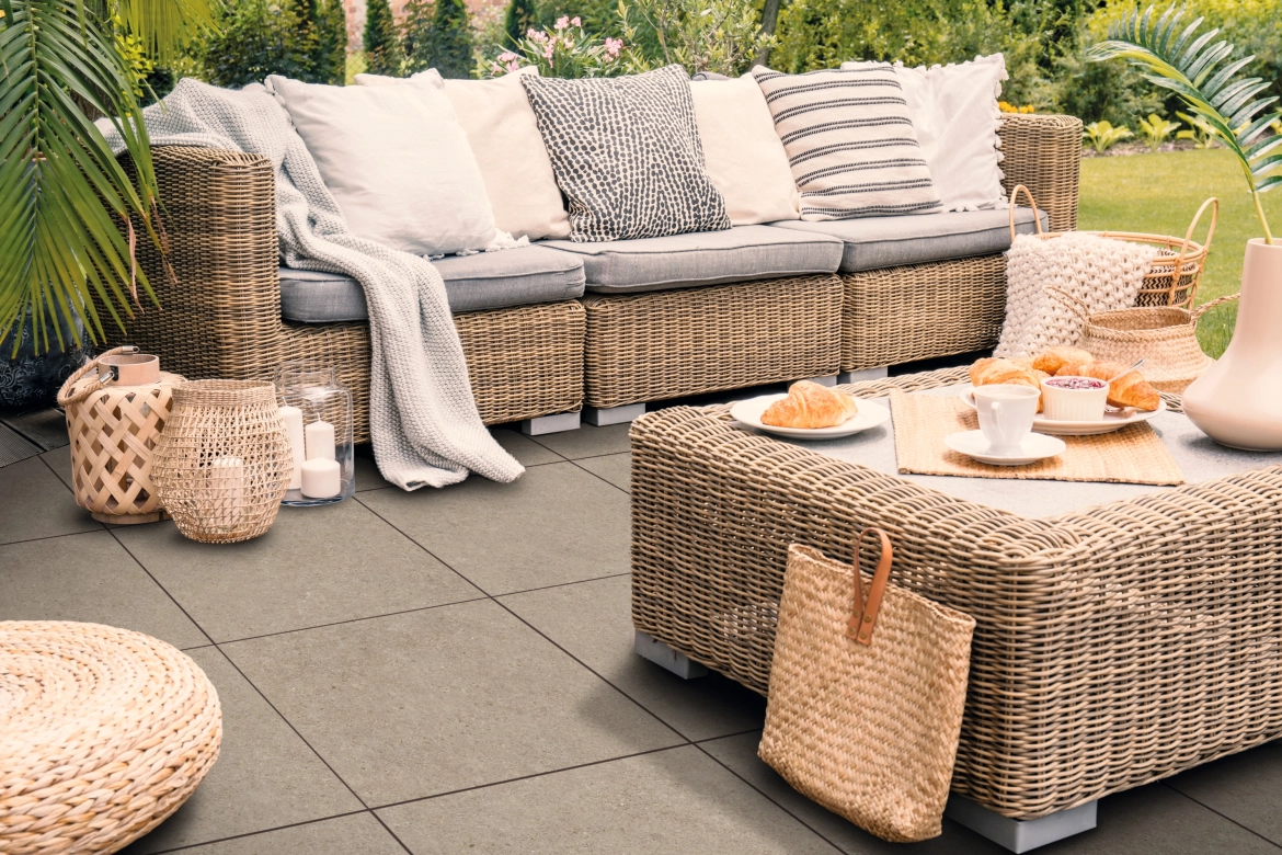 Eine mit Keramikverbundplatten GDM.LIVA twice belegte Terrasse, auf welcher unterschiedliche gewobene Outdoormöbel mit Frühstücksambiente stehen. Im Hintergrund ist ein dicht bewachsener, grüner Garten zu Erkennen.