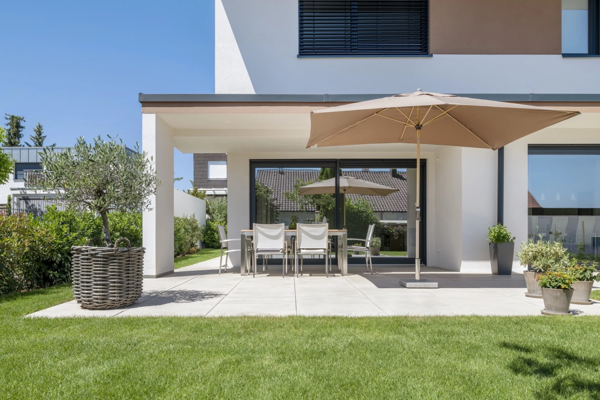 Überdachte Terrasse mit Mobiliar und einem Sonnenschirm. Die terrassenplatten haben eine seidenmatte Haptik und strahlen mit ihrem warmen beigen Farbton Gemütlichkeit und Wärme aus.