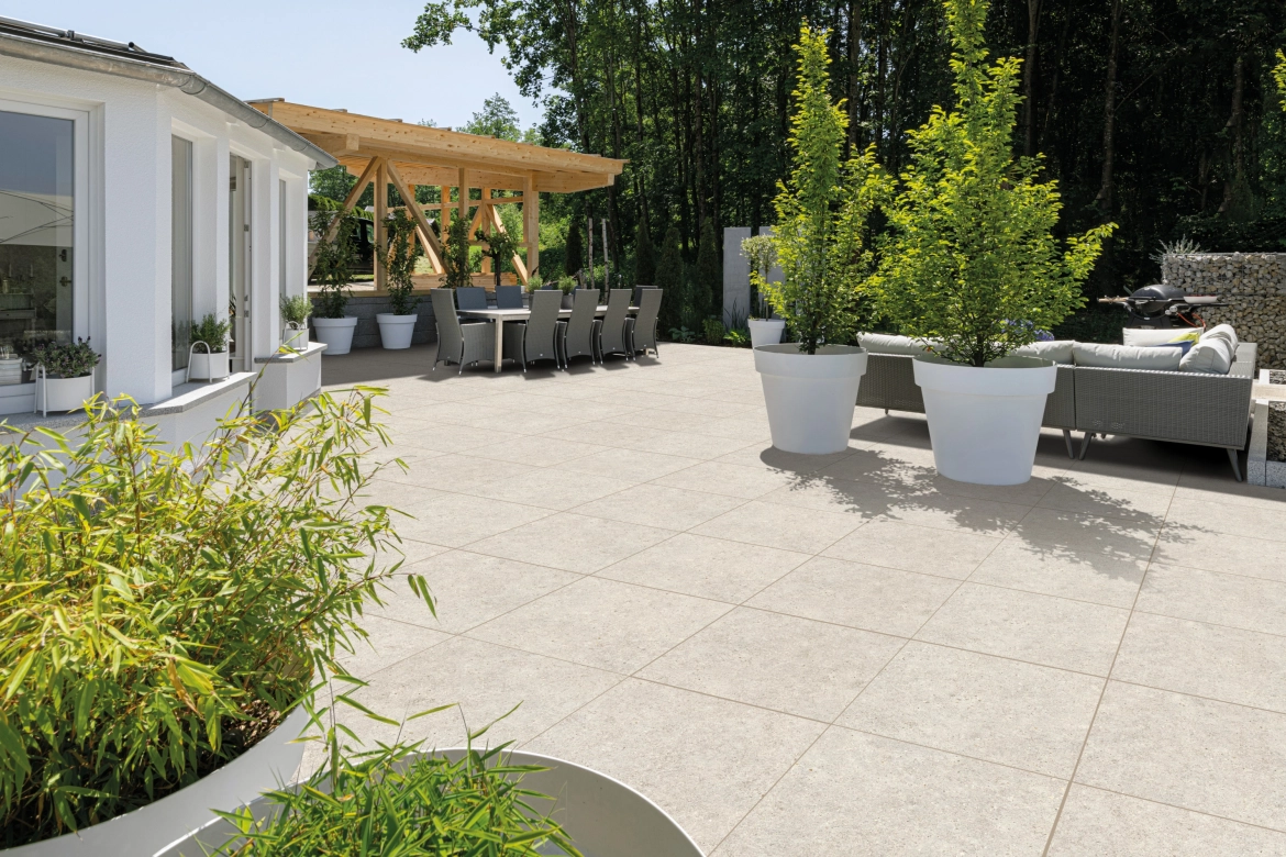 Im Fokus steht eine große Terrasse, auf welcher steingraue Keramikverbundplatten verlegt wurden. Durch verschiedene Outdoormöbel und Pflanzgefäße wird das helle Ambiente auf der Rückseite eines modernen Wohnhauses belebt.