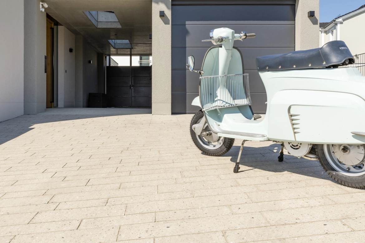 restaurierter, himmelblauer Vespa Roller auf einer beigen Hofeinfahrt, 
belegt mit geschliffenen Pflastersteinen in Terrazzo-Optik in länglichem
 Riegelformat, vor einem sandgrauem Haus mit Anthrazitfarbenem 
Garagentor und abgestimmter Außenbeleuchtung.