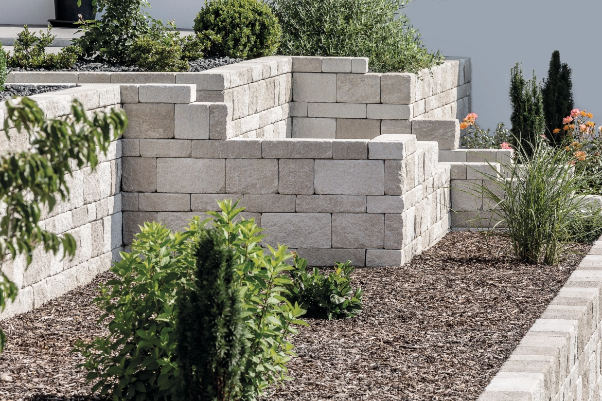 Terrassierter Hang mit einer Mauer aus Naturstein ind sandgrau und 
angelegten Pflanzbeeten geüllt mit Rindenmulch, Schilf und Gräsern.