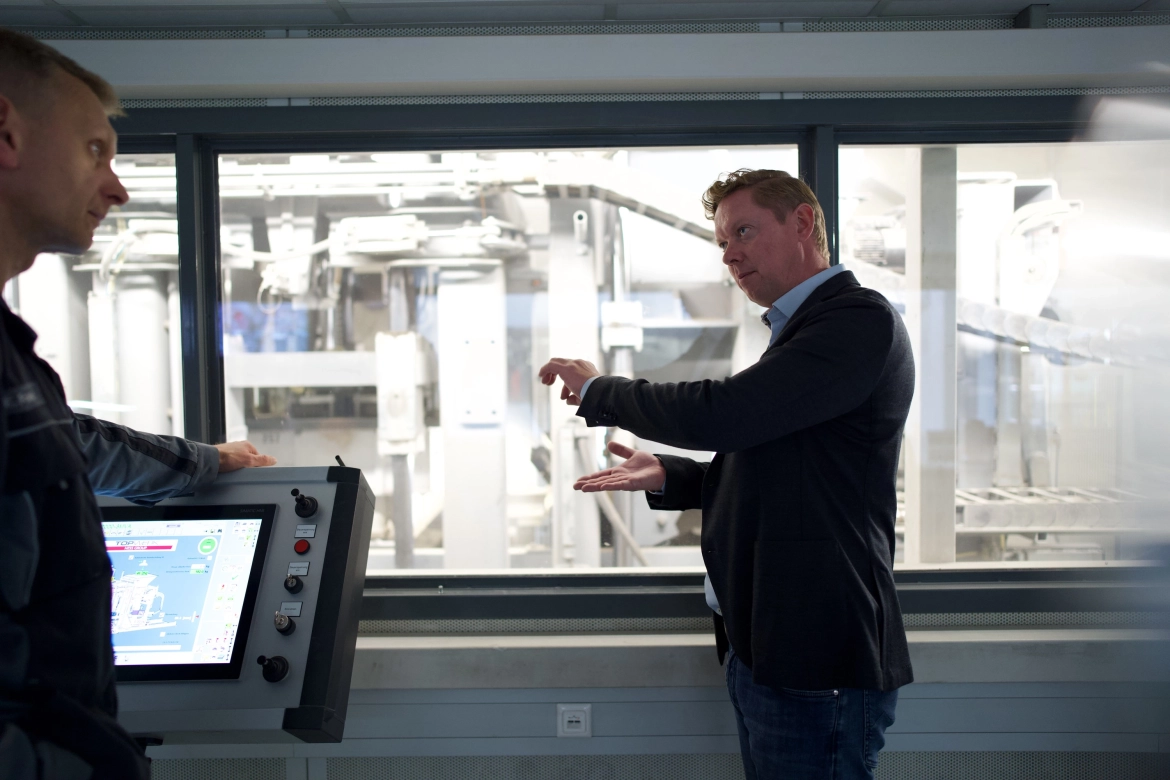 Bernhard Godelmann in conversation with an employee in a machine operator's cabin. In the background, the paving stone machine can be seen.