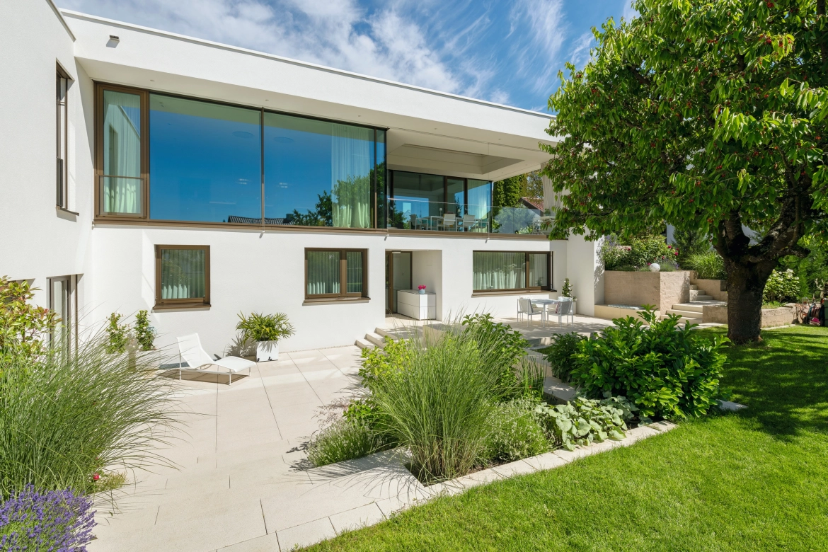 Ein modernes Haus mit Glasfronten und stylischer Terrasse mit üppiger Bepflanzung und hellen, großformatigen Plattenbelägen in Sandstein Farben