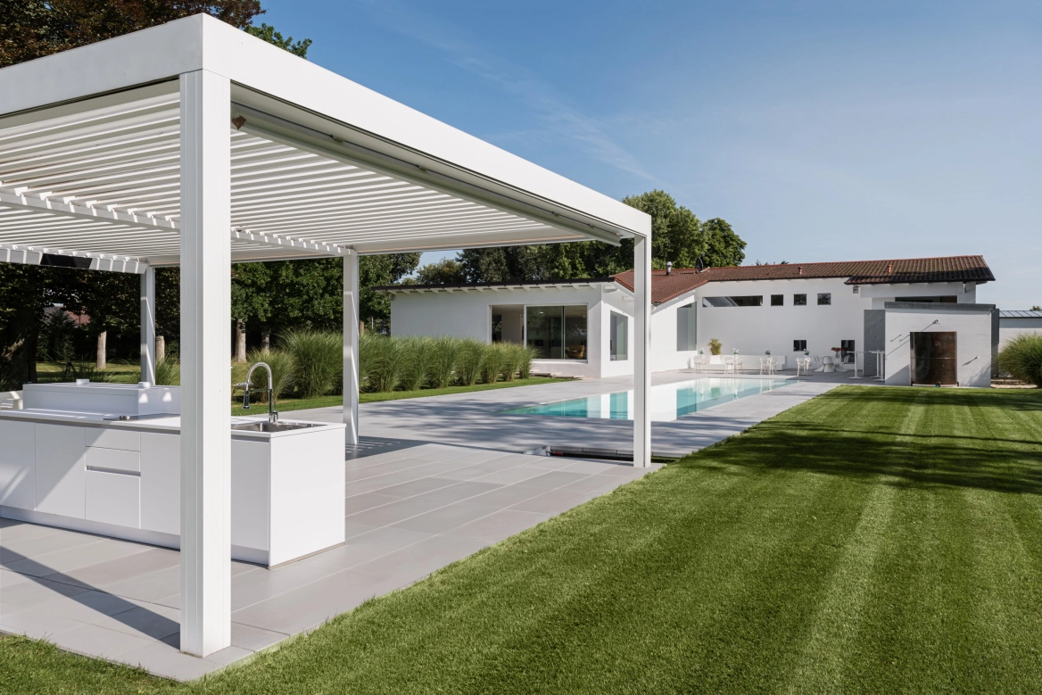 Überdachte Outdoorküche auf länglichen Sichtbetonplatten in grau vor einem Pool mit Einfassung aus Beton und angrenzendem Rasen.