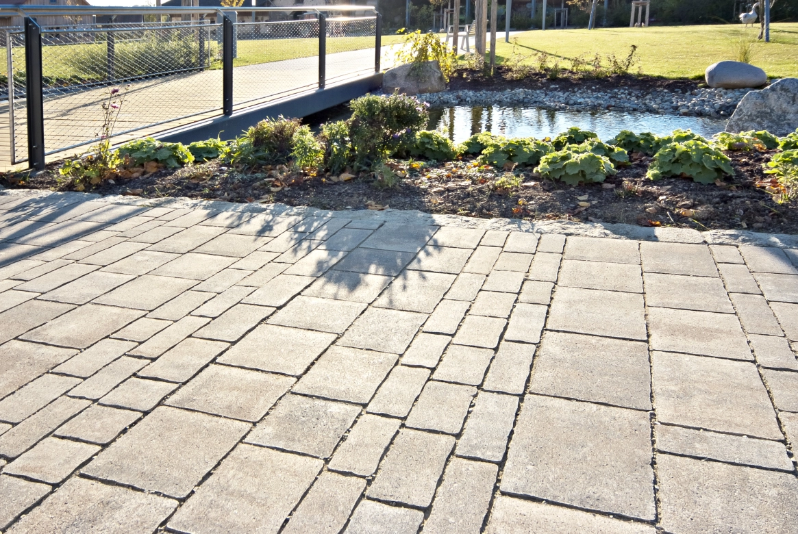 gepflasterter Weg mit klein- und großformatigen PFlastersteinen im 
Reihenverband mit geschwungenen Kanten. Die Umrandung des Weges besteht 
aus bepflanzten Beeten und einem angelgeten Bachlauf über den ine Brücke
 führt.
