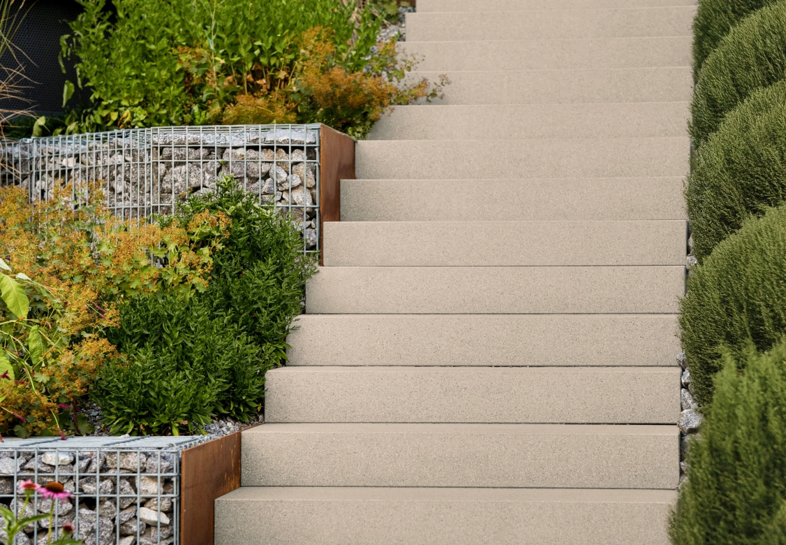 Kalksteinfarbene Treppe bestehend aus den Stufen GDM.LIVA step, welche den Hang eines mit Pflanzen, Streuchern und Hecken bepflanzten Hangs im Garten hinaufführt