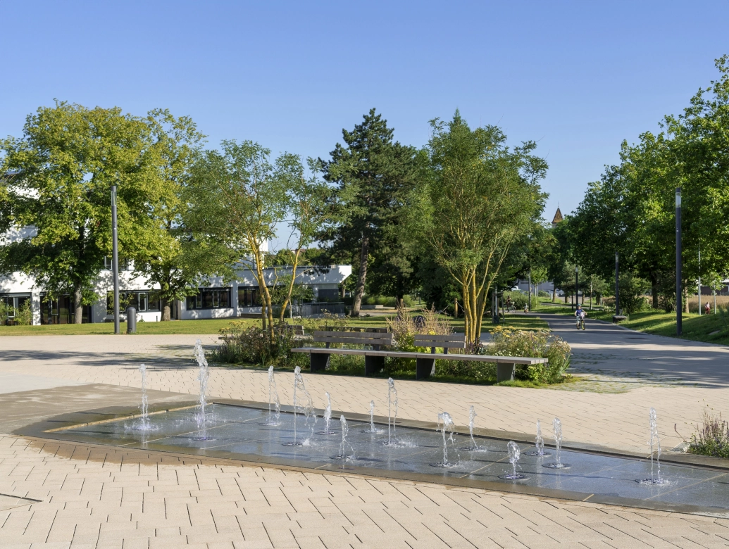 Offener Platz in einer begrünten Park-Umgebung. Inmitten des gepflasterten Platzes ist ein Wasserspiel mit Springbrunnen Elementen platziert. Im Hintergrund sind viele Grünflächen mit Wiesen, Sträuchern und Bäumen. Dahinter ist ein Gebäude mit weißer Fassade zu erkennen.