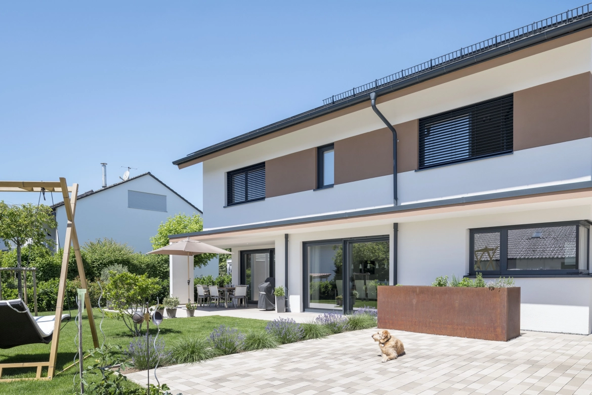 Garden with terrace and grassed area and paved area where a dog sits.