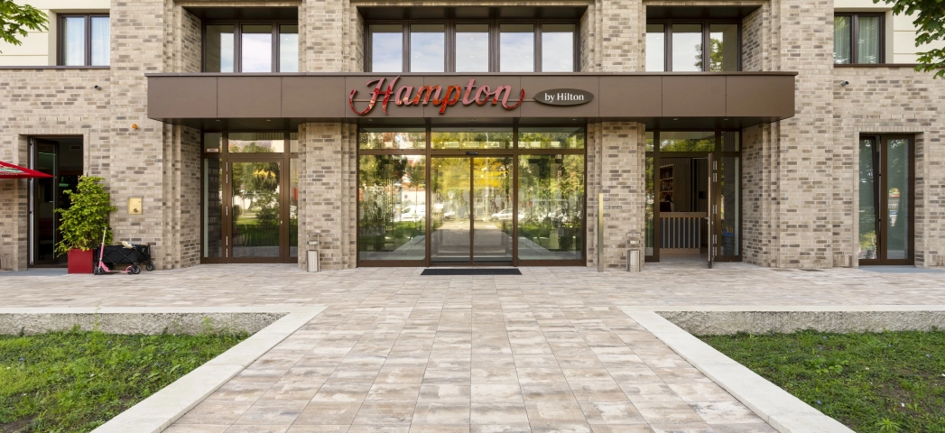 Moderne Gebäudefassade mit braunen Wandfliesen, vielen großen Fenstern und dunkelbraunem Schild über dem Eingang. Davor ist ein gepflasterter Weg aus muschelkalkfarbenen Pflastersteinen - dem GDM.KLIMASTEIN. Daneben befinden sich Grünflächen.