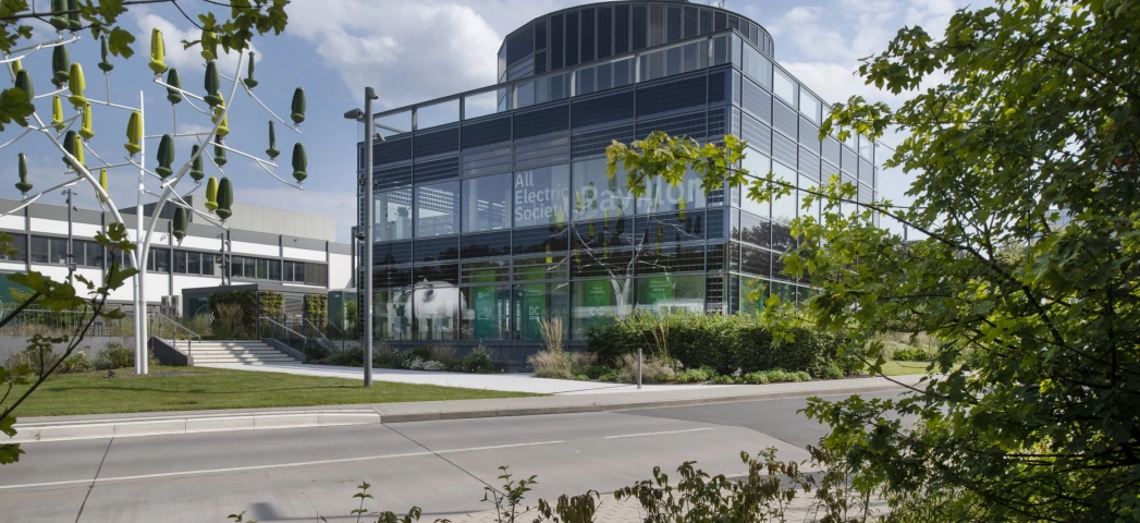 Ein modernes Gebäude mit einer grün-bläulichen Fenster-Fassade. Im Vordergrund ist eine Straße, Pflanzen, und eine Skulptur, die an einen Baum erinnert zu erkennen.