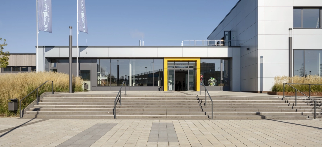Großer Vorplatz mit grauem GDM.SCENIC individual und dunkleren Betonsteinen zur gestalterischen Auflockerung. Eine Treppe führt auf das große Gebäude mit grauer Fassade zu. Um den Eingangsbereich herum ist eine auffällige gelbe Umrandung.