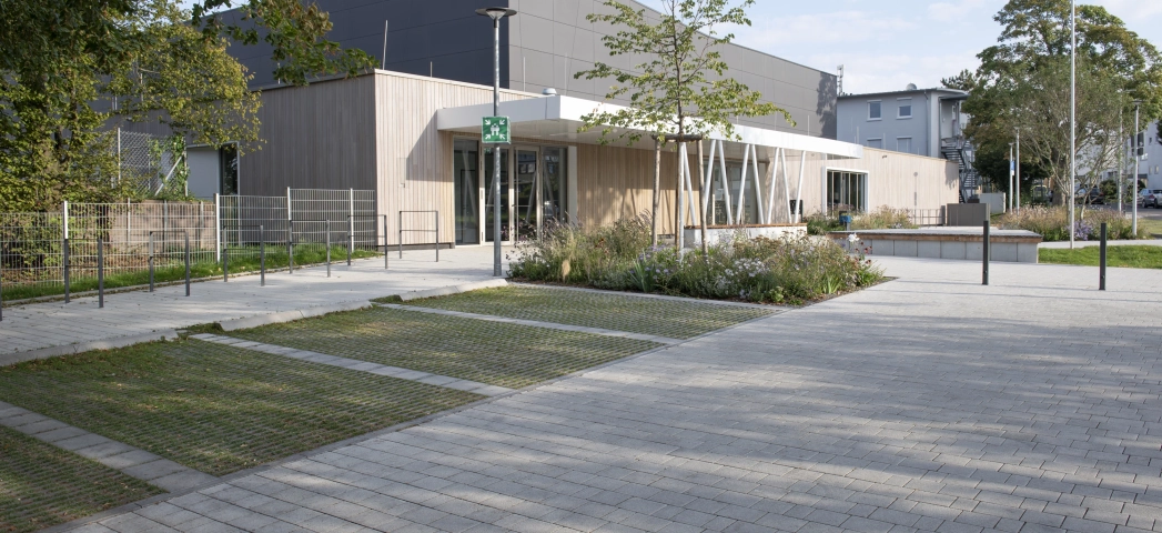 Moderne Außenfassade einer Sporthalle mit anthrazitfarbenen Platten im oberen Bereich und Holzverkleidung im unteren Bereich. Davor befindet sich ein mit Kalk-Grauen, versickerungsfähigen Betonsteinen gepflasterter Vorplatz mit Parkplätzen aus Rasengittersteinen und Sträuchern, Gräsern und Bäumen. 