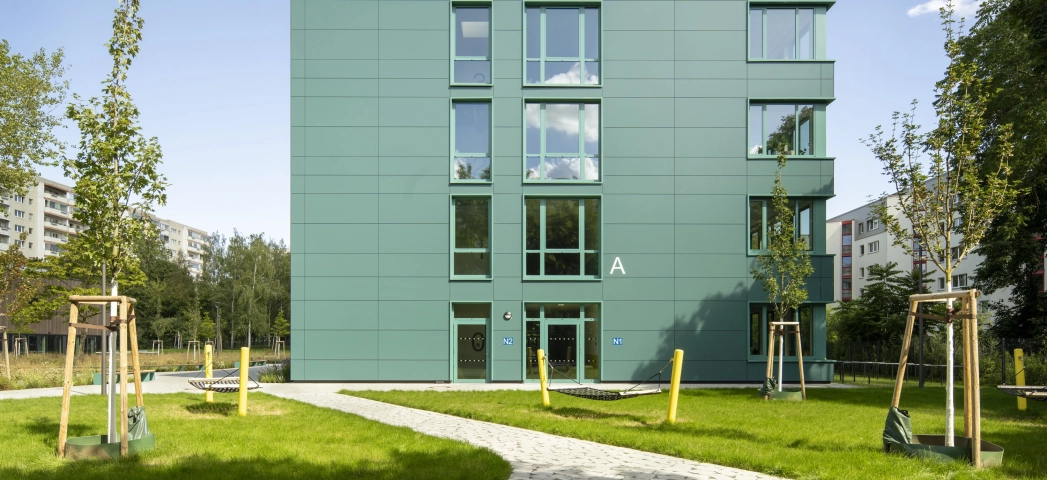 Ein mit rundlich gepflasterteten Betonsteinen gepflasterter Weg, welcher direkt zu einem modernen Schulgebäude mit petrolfarbener Fassade führt. Der Bereich links und rechts neben dem Pfad ist mit Streuchern, Wiesen und Bäumen begrünt.