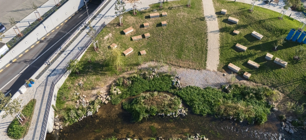 Luftaufnahme eines neu gestalteten Parkareals mit gepflasterten Wegen, einem kleinen Bachlauf, Grünflächen mit vielen Sitzbänken sowie angrenzenden Spiel- und Verkehrsbereichen.