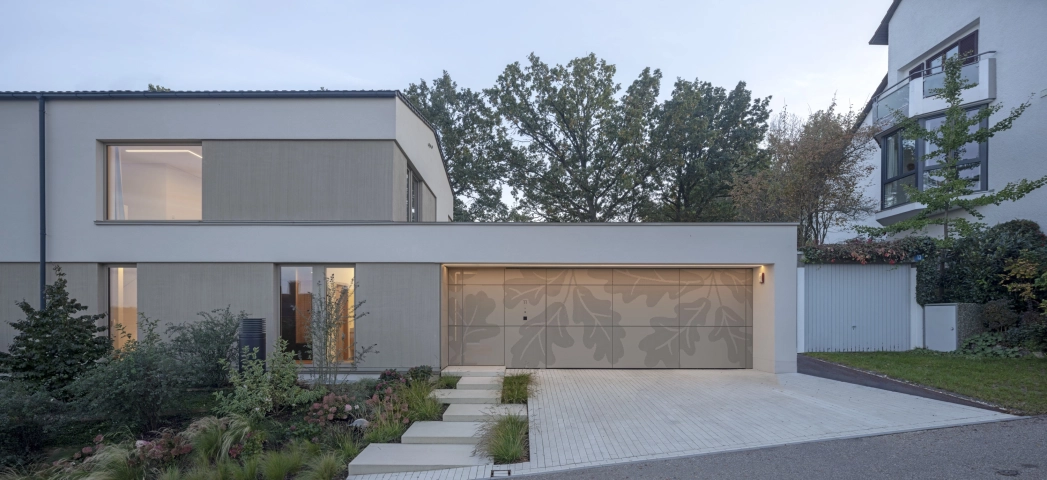 Abendliche Aufnahme eines hell beleuchteten, modernen Wohnhauses. Im Vordergrund ist eine gepflegte Einfahrt mit indivdualisierten Garagentoren und ein wild bepflanztes Blumenbeet zu erkennen.