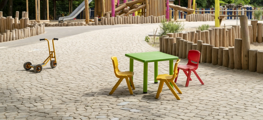 Grüner Kindertisch mit zwei gelben und einem roten Stuhl auf GDM.BOLERO. Links ein gelber Kinderroller, dahinter Spielgeräte aus Naturholz.