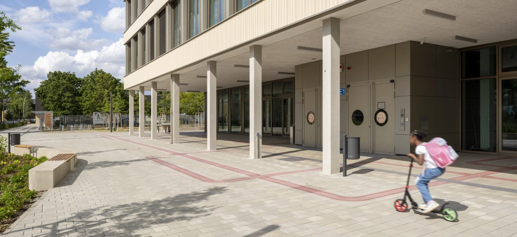 Fassade eines Schulgebäudes mit großen Fenstern und Säulen. Davor liegt ein grau-beigen geplasterter Platz mit Sitzelementen und einem Kind, welches mit einem Roller fährt. Im Hintergrund sind viele Bäumen zu erkennen.