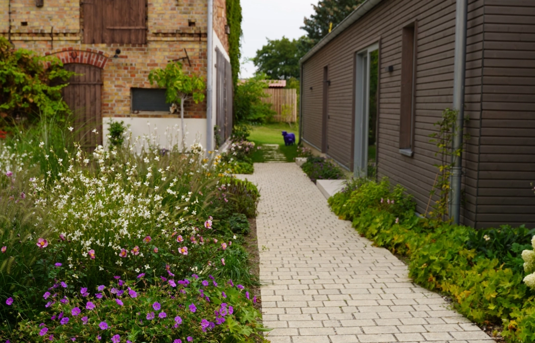 Ein rustikales Backsteingebäude mit großen Holztoren und einen angelegten Garten im Vordergrund. Ein gepflasterter Weg führt durch den Garten, der mit Blumen und Sträuchern bepflanzt ist. Vor dem Garten ist ein naturbeige gepflasterter Hof im Reihenverband angelegt auf welchem ein Kind auf dem Fahrrad fährt. Rechts ist ein modernes Gebäude mit großen Glastüren zu sehen.