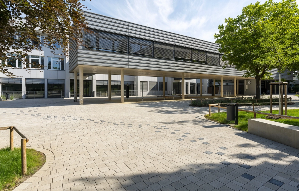Offener Schulhof umgeben von grünen Wiesen, Bäumen und Sitzelementen. Ein Pfad bestehend aus anthrazitfarbenen Betonsteinen zieht sich von rechts nach links durch den gepflastertet Hof bis hin zu einem modernen, mehrteiligen Schulgebäude. Der vordere Teil steht auf Betonsäulen und ist dunkelgrau verkleidet. Der hintere Teil hat eine weiße Fassade. 