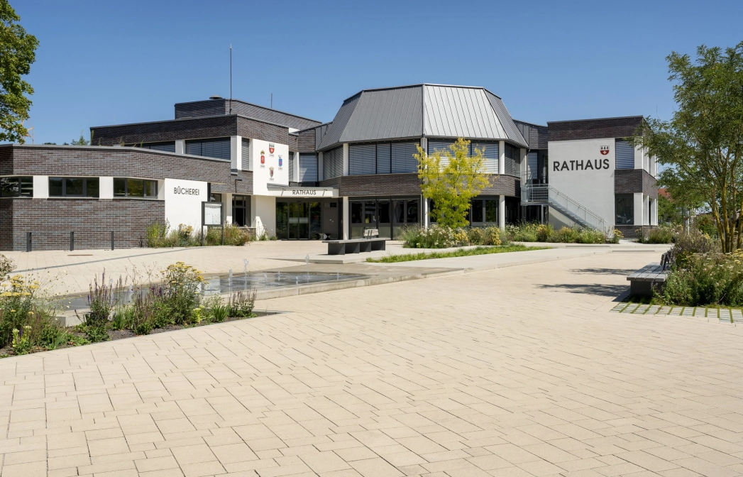 Offener Platz in einer städtischen Umgebung. Inmitten des gepflasterten Platzes ist ein Wasserspiel mit Springbrunnen Elementen platziert , welches von Blumenbeeten und Bäumen umrahmt wird. Im Hintergrund ist ein modernes Rathaus zu sehen, das vom  Platz umschlossen wird.