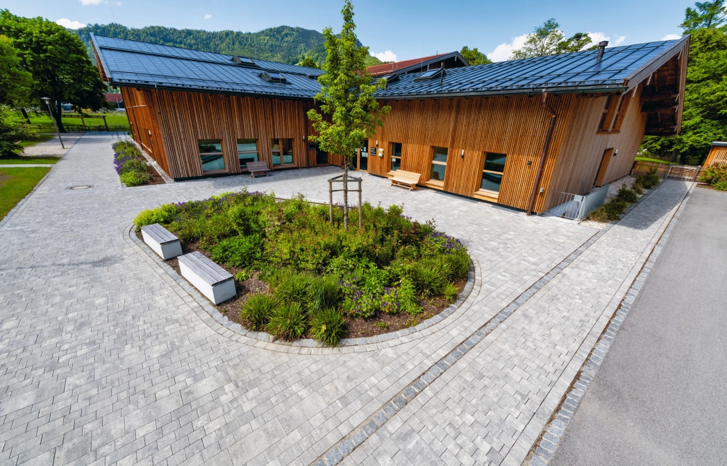 Modern gepflasterter Platz mit Grünfläche und Sitzelementen vor einem Bürgerzentrum  mit Holzfassade