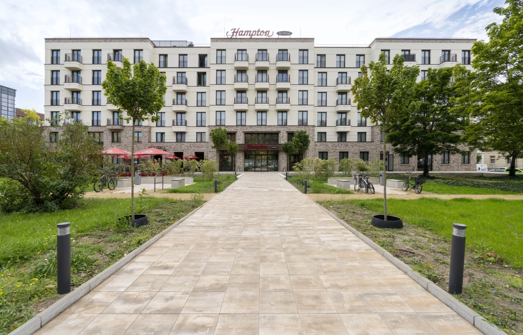 Blick auf großes Hotelgebäude mit vielen Fenstern, beiger Fassade im oberen Bereich und braunen Wandfiesen im unteren Bereich. Davor ist ein Weg aus muschelkalkfarbenem GDM.KLIMASTEIN. Drumherum sind Grünflächen, Bäume und Sträucher.