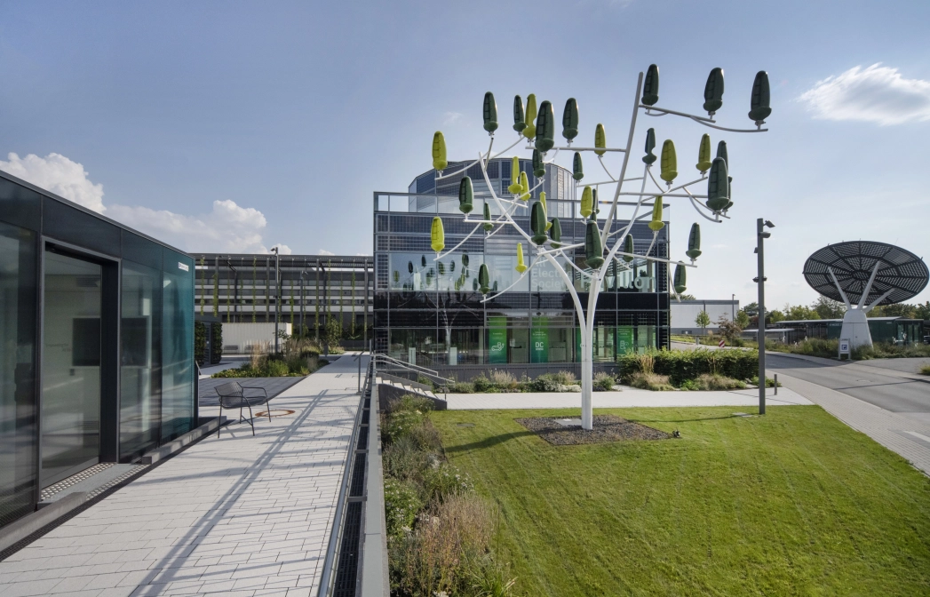 Ein modernes Gebäude mit einer grün-bläulichen Fenster-Fassade. Im Vordergrund ist eine nachhaltig gestaltete Pflasterfläche, eine Wiese und eine Skulptur, die an einen Baum erinnert zu erkennen. Im Hintergrund ist eine große Satelliten-Schüssel zu sehen.