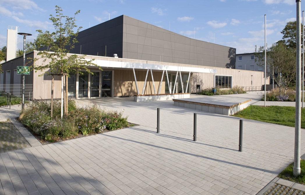 Moderne Außenfassade einer Sporthalle mit anthrazitfarbenen Platten im oberen und Holzverkleidung im unteren Bereich. Davor befindet sich ein mit Kalk-Grauen, versickerungsfähigen Betonsteinen gepflasterter Vorplatz mit Sträuchern, Gräsern und einem kleinen Baum.