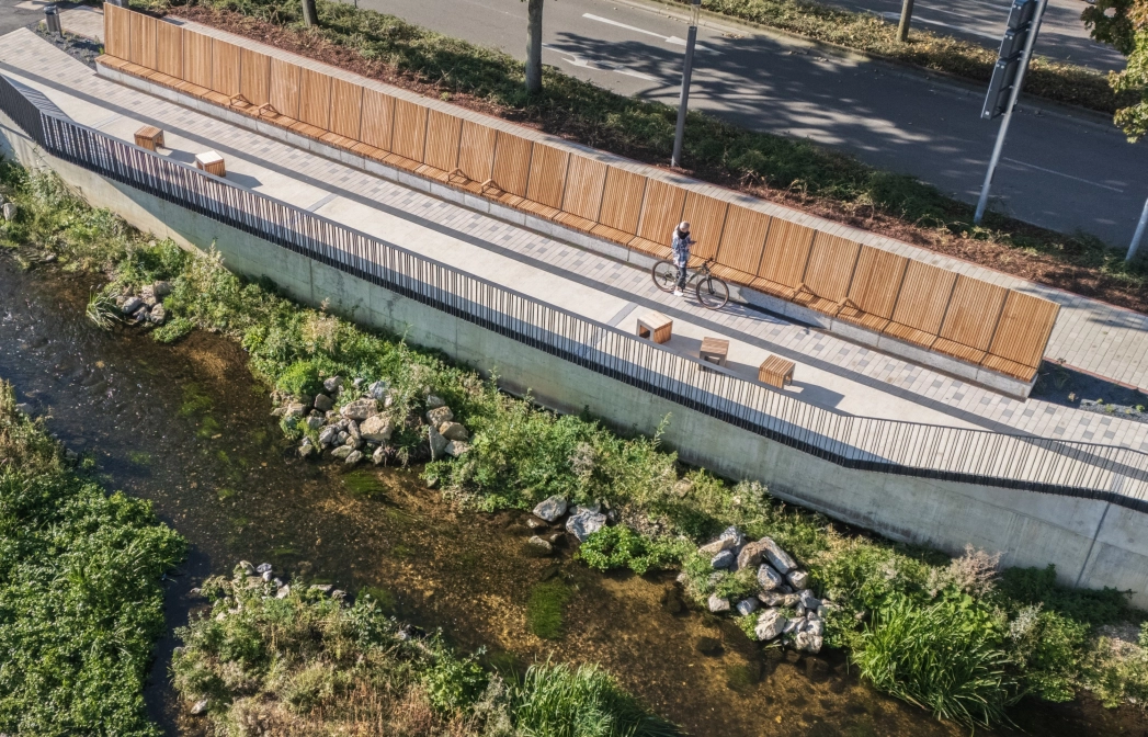 Luftaufnahme eines Uferwegs entlang eines flachen Bachlaufs, gesäumt von Holzsitzbänken und einer langen Holzverkleidung. Der Weg ist mit hellem Betonstein gepflastert, während oberhalb eine Straße mit Bäumen verläuft.