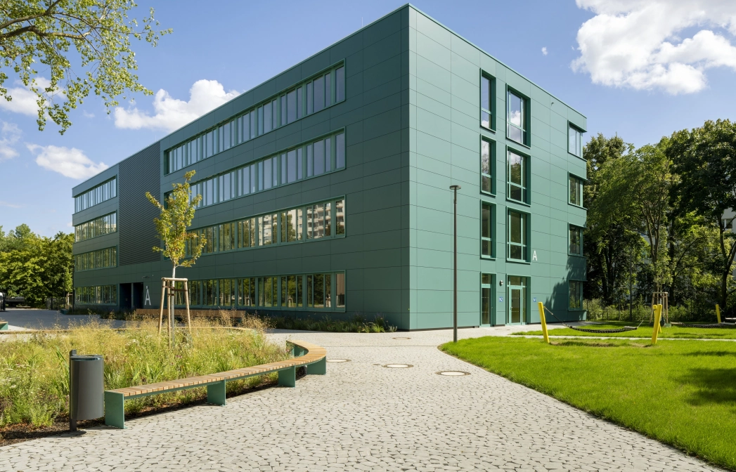 Ein mit rundlich gepflasterteten Betonsteinen gepflasterter Weg, welcher direkt zu einem modernen Schulgebäude mit petrolfarbener Fassade führt. Der Bereich links und rechts neben dem Pfad ist mit Streuchern, Wiesen und Bäumen begrünt.