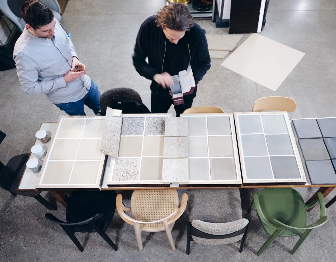 Square color samples lie on a table, ranging from light beige tones to dark anthracite colors. Two people are bent over the color samples, examining the colors.