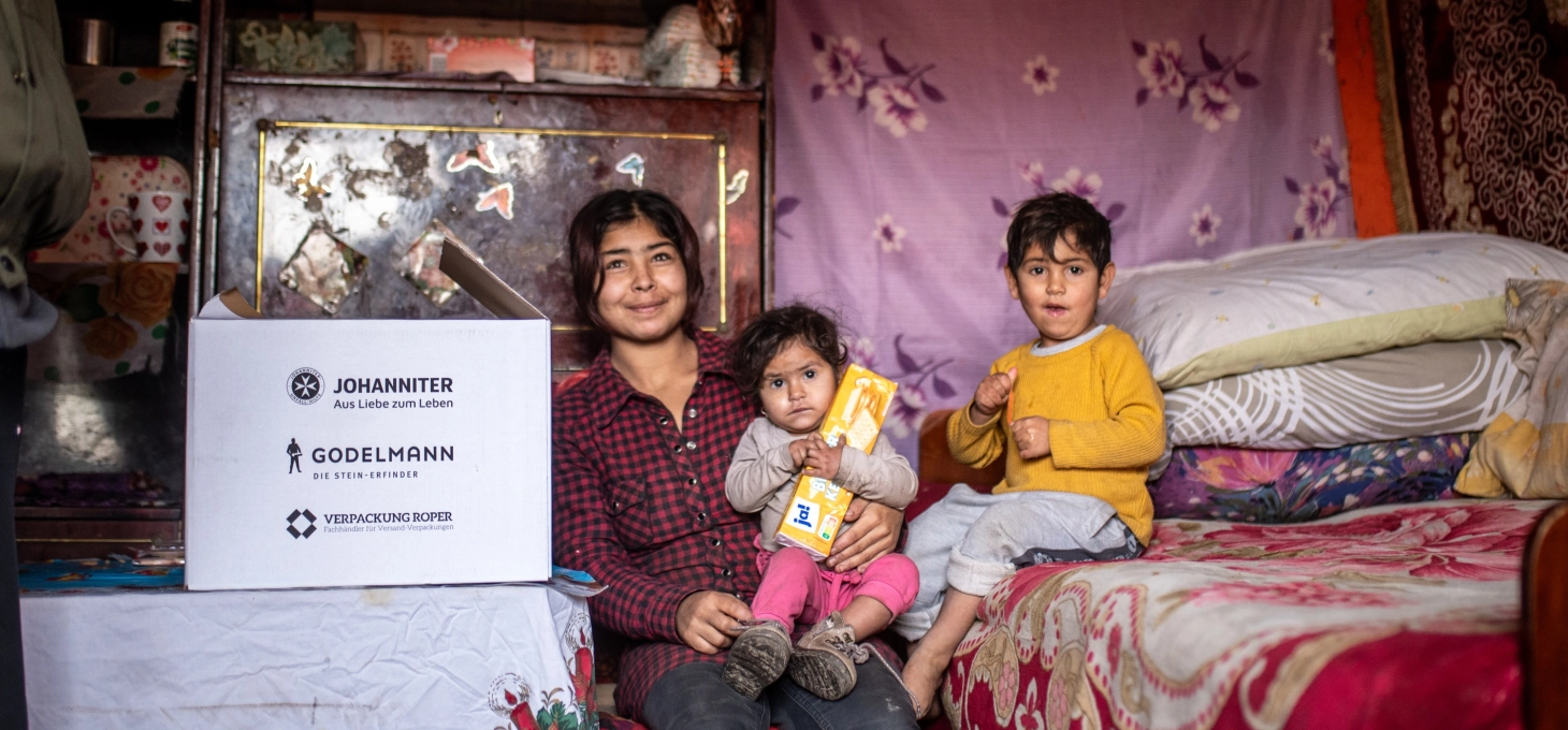 Hilfe vor Ort: GODELMANN Spenden-Päckchen steht auf einem Tisch, daneben sitzt eine rumänische Familie bestehend aus einer Mutter mit ihren zwei Kindern.