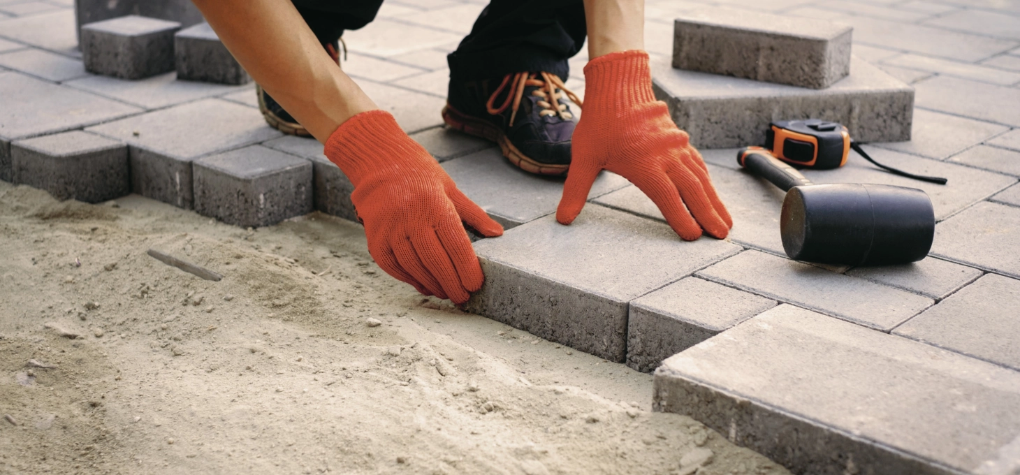 Eine Person mit roten Handschuhen verlegt graue Pflastersteine in eine Fläche. Neben ihm liegt Werkzeug, unter anderem ein Maßband und ein Gummihammer.