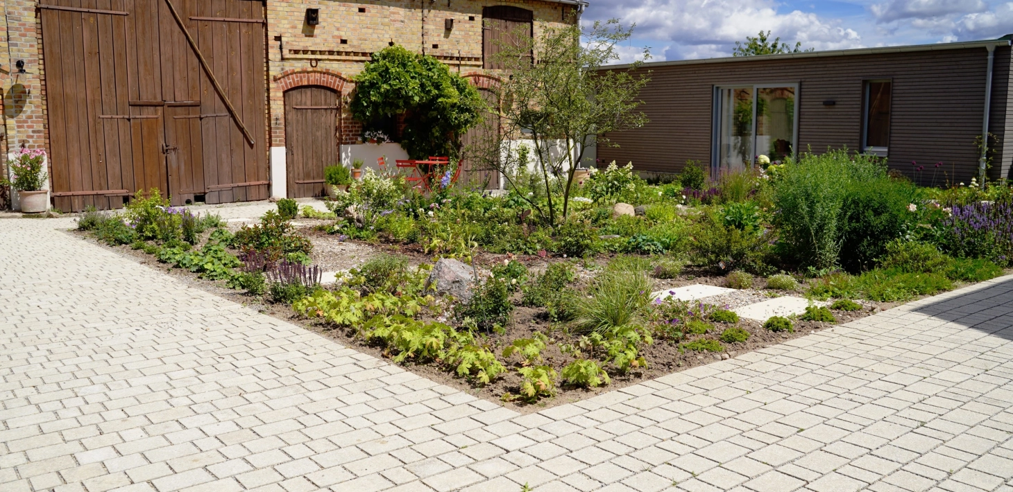 Ein rustikales Backsteingebäude mit großen Holztoren und einen angelegten Garten im Vordergrund. Ein gepflasterter Weg führt durch den Garten, der mit Blumen und Sträuchern bepflanzt ist. Vor dem Garten ist ein naturbeige gepflasterter Hof im Reihenverband angelegt. Rechts ist ein modernes Gebäude mit großen Glastüren zu sehen.