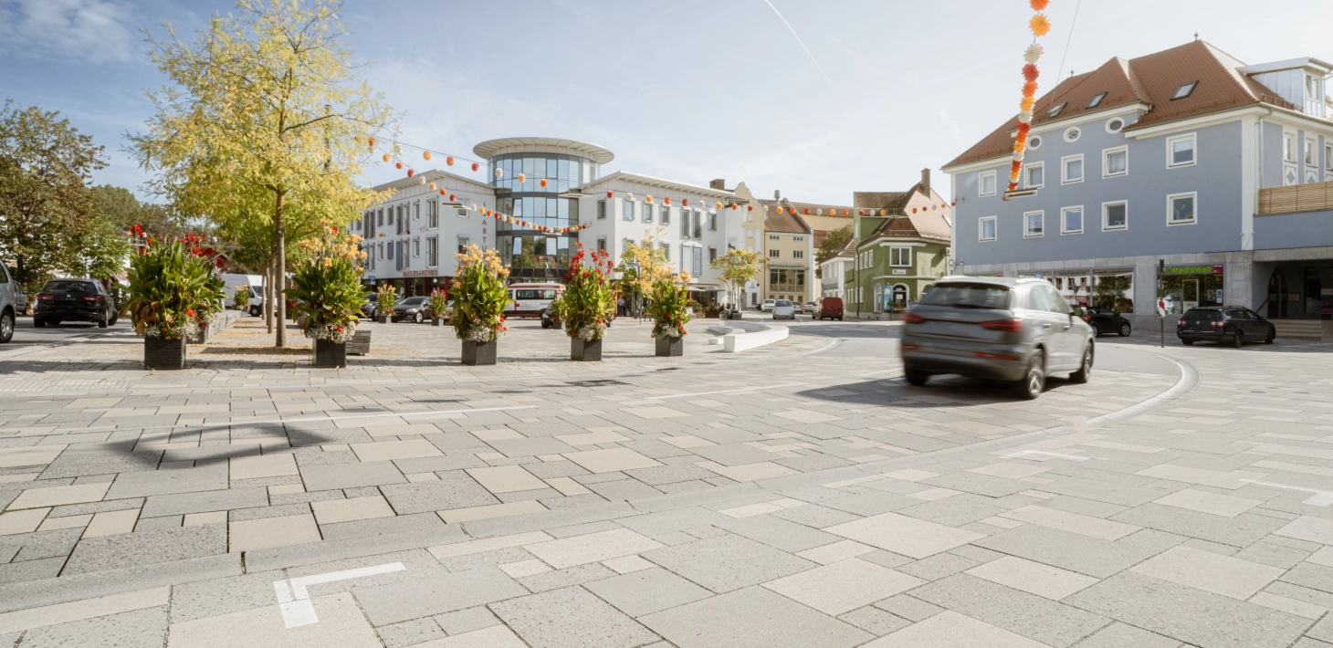 befahrbare Großfromatplatten im wilden Verband und abgestuften Grautönen auf einem Stadtplatz mit Bepflanzung.