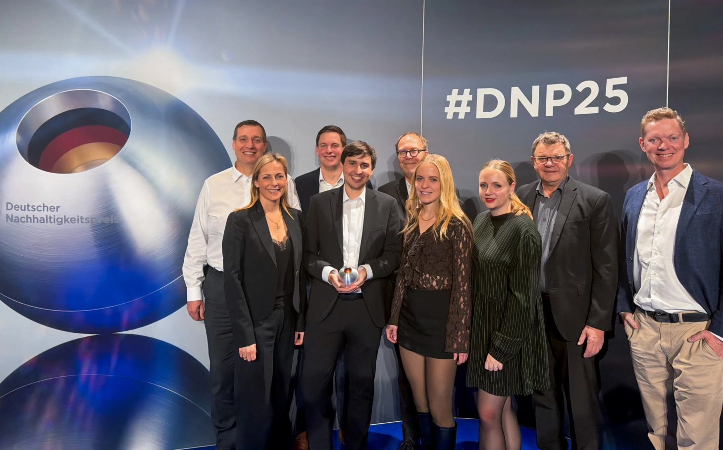 Gruppenfoto mit 9 Personen vor einer Fotowand auf der der Deutsche Nachhaltigkeitspreis abgebildet ist. Die Godelmann-Gruppe freut sich über den Deutschen Nachhaltigkeitspreis für den GDM.KLIMASTEIN