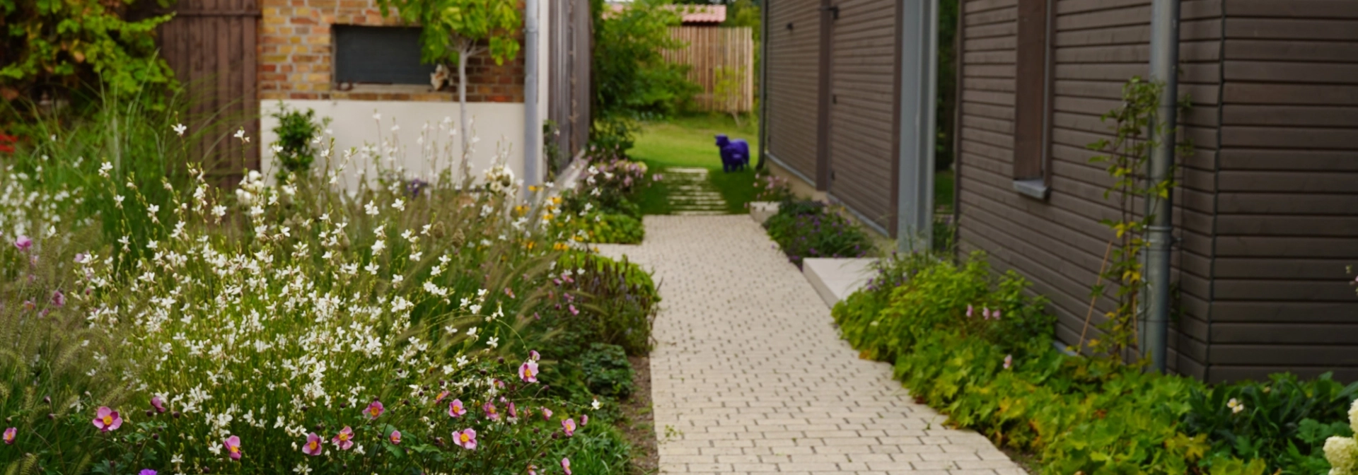 Ein rustikales Backsteingebäude mit großen Holztoren und einen angelegten Garten im Vordergrund. Ein gepflasterter Weg führt durch den Garten, der mit Blumen und Sträuchern bepflanzt ist. Vor dem Garten ist ein naturbeige gepflasterter Hof im Reihenverband angelegt auf welchem ein Kind auf dem Fahrrad fährt. Rechts ist ein modernes Gebäude mit großen Glastüren zu sehen.