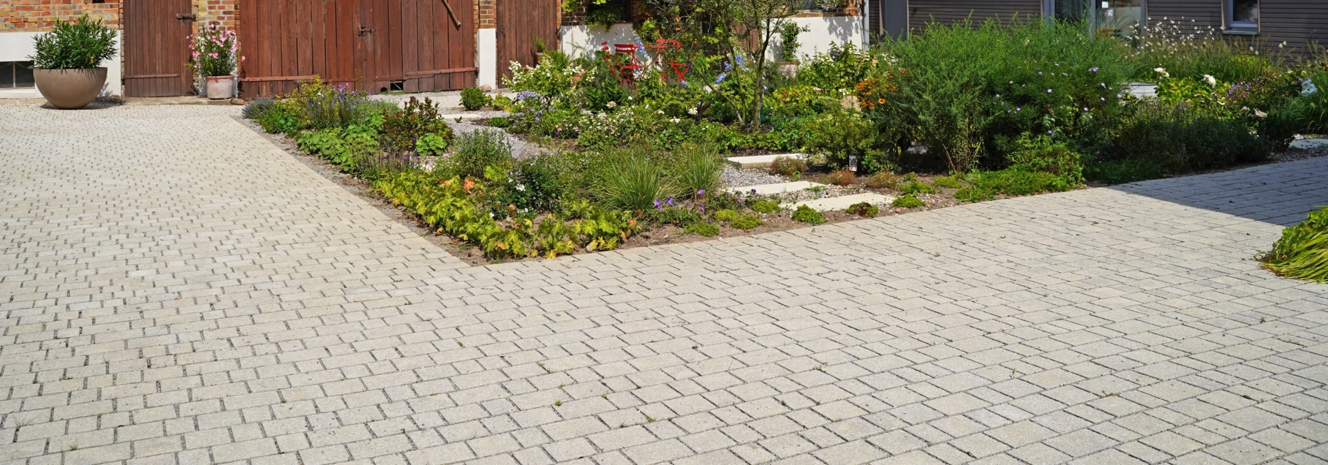 Ein rustikales Backsteingebäude mit großen Holztoren und einen angelegten Garten im Vordergrund. Ein gepflasterter Weg führt durch den Garten, der mit Blumen und Sträuchern bepflanzt ist. Vor dem Garten ist ein naturbeige gepflasterter Hof im Reihenverband angelegt. Rechts ist ein modernes Gebäude mit großen Glastüren zu sehen.