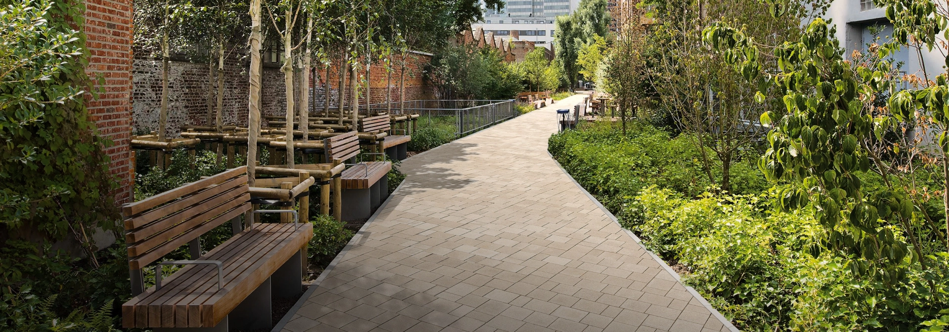 A path paved with evaporative concrete blocks leads straight through a leafy park in the middle of a large city. The paving stones are shaded beige and have a bead-blasted surface. To the left along the path is a brick wall, in front of which there are several benches and trees. To the right are more small trees, shrubs and grasses, behind which a brick house is hidden. High-rise buildings can be seen in the background.  Translated with DeepL.com (free version)