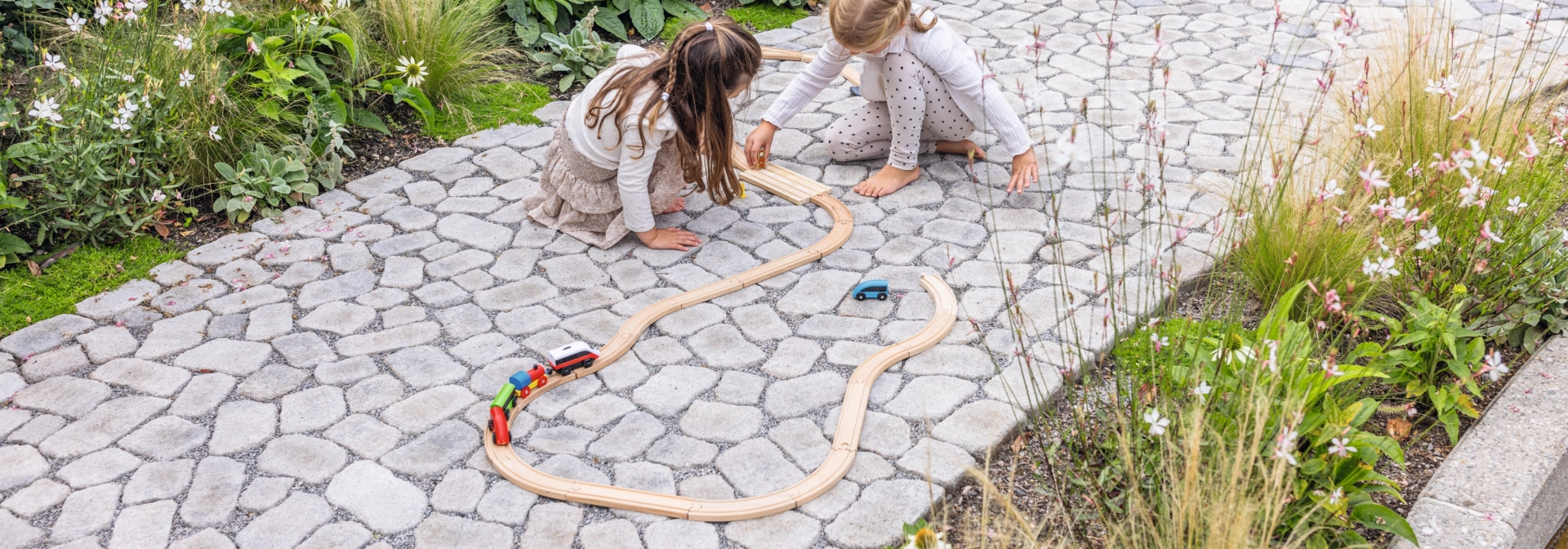 Zwei Kinder spielen mit einem Holzzug auf einem gepflasterten Gartenweg.