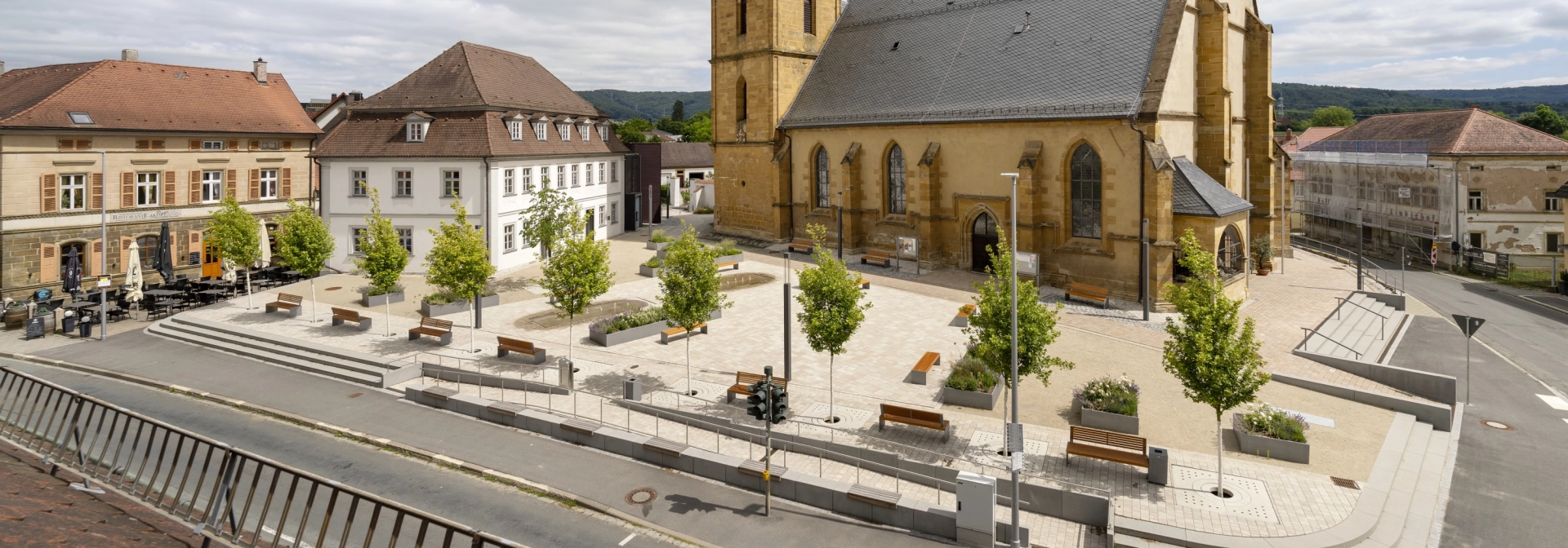 Eine Totale des Kirchenumfelds der St. Kilian Kirche in Scheßlitz. Im Vordergrund ist ein heller Platz mit Bäumen und Wasserbecken und im Hintergrund eine große Kirche zu sehen.