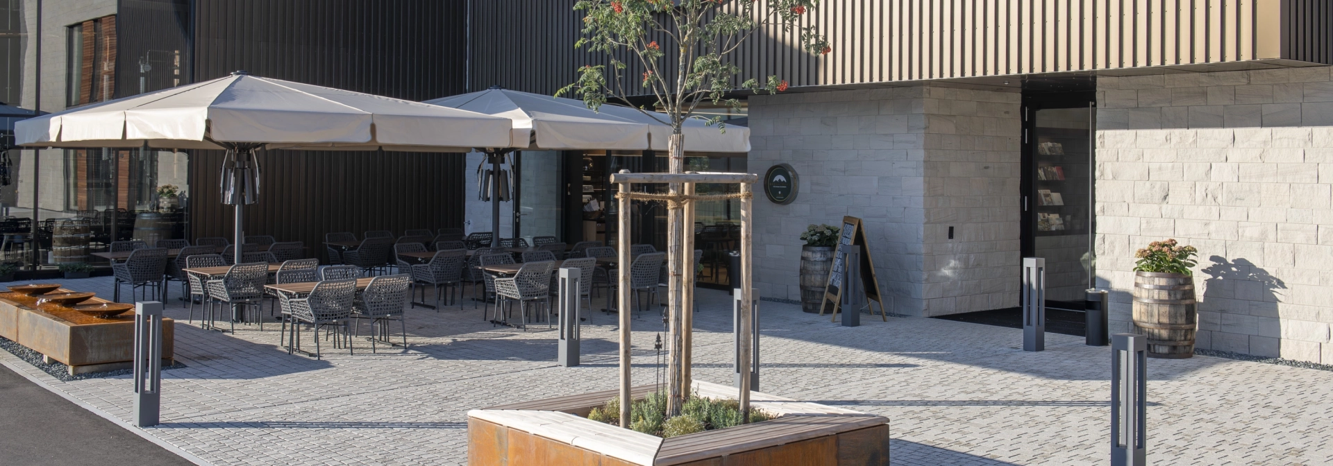Moderner, gepflasterter Außenbereich mit Tischen und Stühlen unter weißen Sonnenschirmen vor einem modernem Gebäude mit Holz- und Steinfassade. Im Vordergrund ist ein kleinerer Baum, mit einer Einfassung aus rostigem Corthenstahl und Holzsitzfläche.