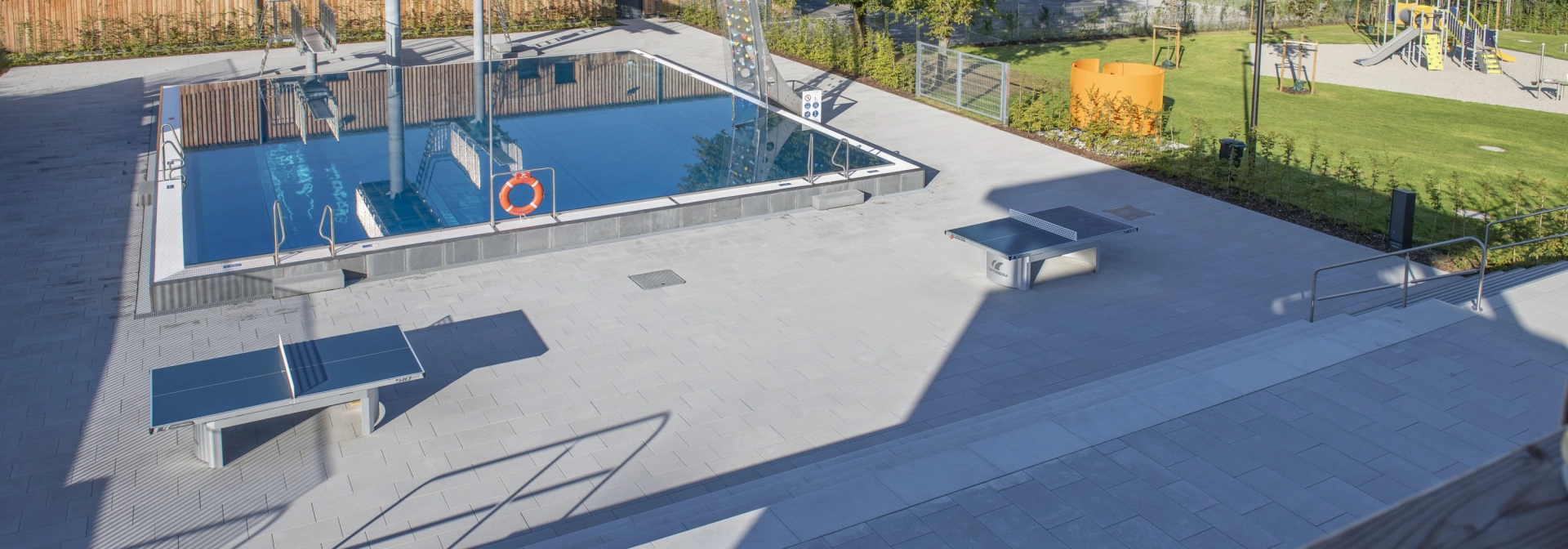Modernes Freibad mit rechteckigem Edelstahlbecken und weitläufiger, heller Terrasse mit mehreren Tischtennisplatten; im Hintergrund liegen eine Rasenfläche mit Bäumen und ein Spielplatz.